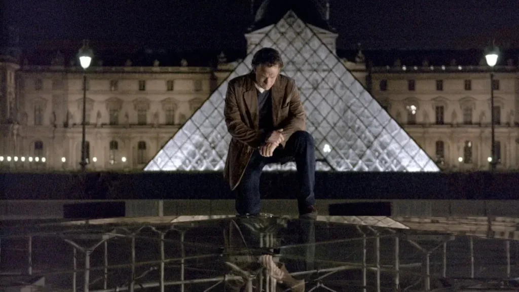Tom Hanks as Robert Langdon kneeling over the glass inverted pyramid outside the illuminated Louvre Pyramid at night in The Da Vinci Code.
