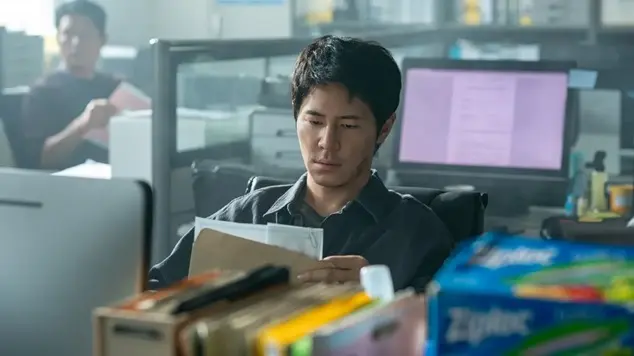 A focused man seated at his office desk examines documents taken from a brown envelope.