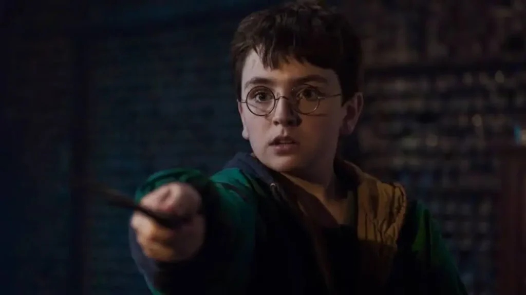 Harry Potter: 3 Things We Loved About the New Series Trailer A close-up of a young boy wearing round glasses and a green jacket, pointing a magic wand directly forward with an intense expression in a dimly lit room.