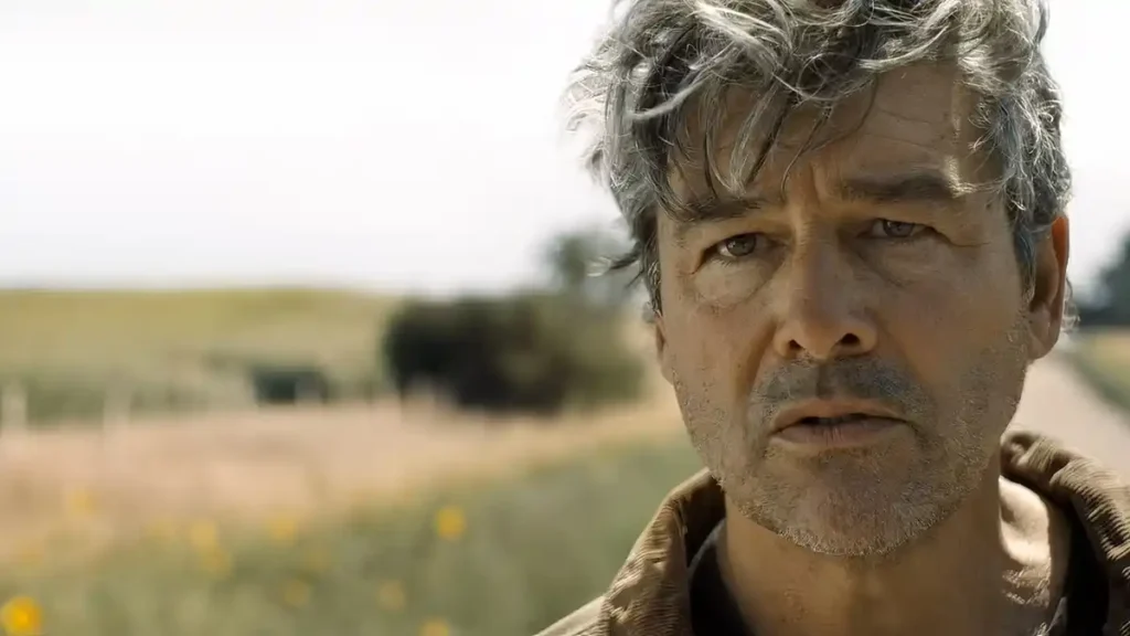 Lanterns Episode 5 Delivers the Season’s Biggest Twist Yet Close-up of a middle-aged man with tousled grey hair and stubble, looking intently and slightly troubled past the camera against an out-of-focus rural field and road.